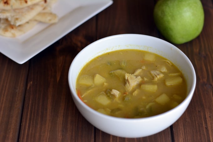 A white bowl filled with Indian mulligatawny soup next to a green apple and a white plate with Naan bread.