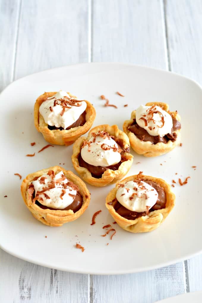 Five mini chocolate cream pies topped with whipped cream and chocolate shavings on a white plate.