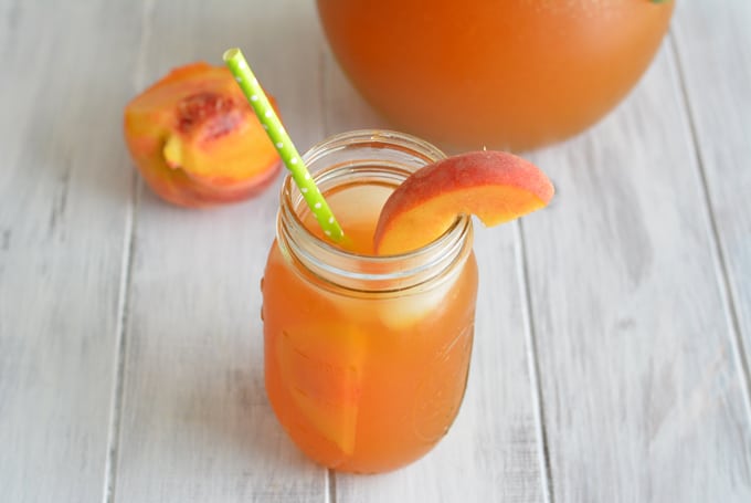 A mason jar filled with peach iced tea with a peach slice and green straw.