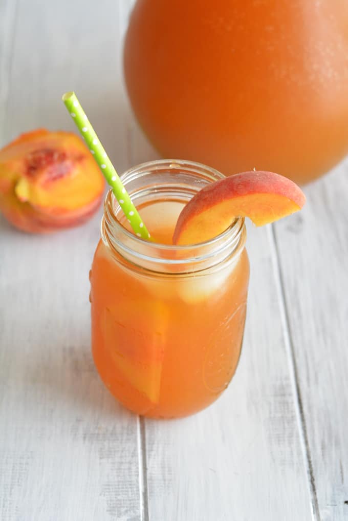 Peach flavored green iced tea in a mason jar with a peach slice and green straw with white polka dots.