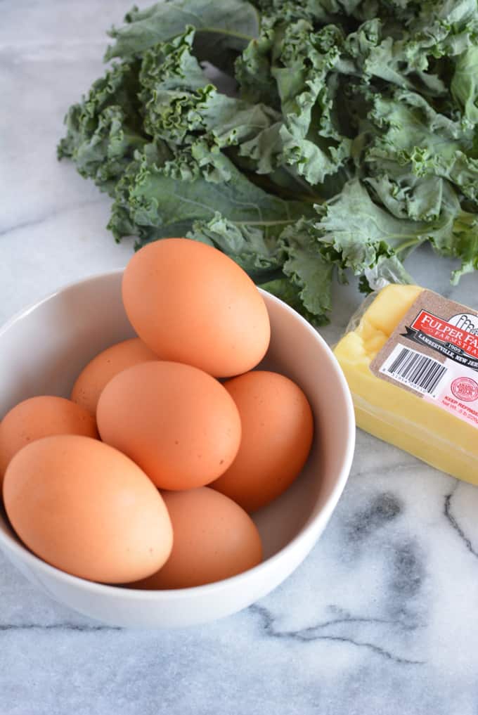 Brown eggs with a bunch of kale and block of cheese on a white marble table.