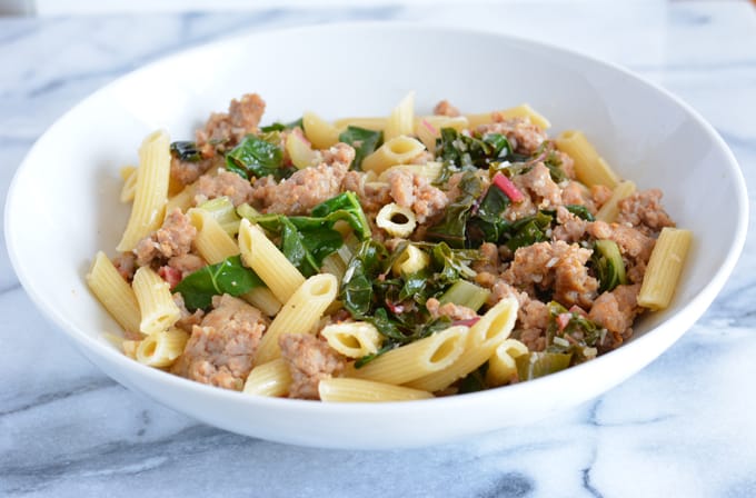 A white bowl filled with penne pasta, Swiss chard and sausage.