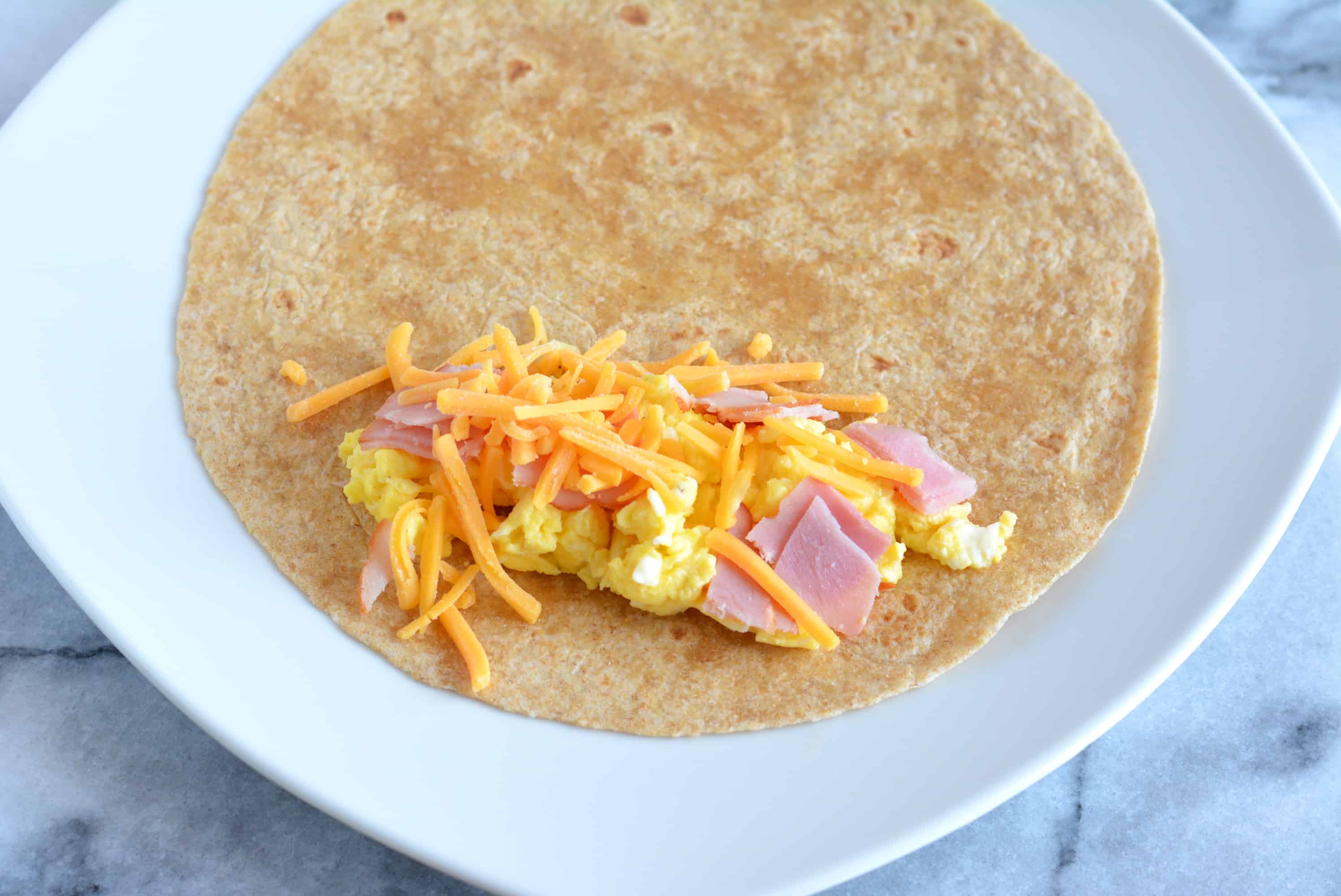 Scrambled eggs, shredded cheddar cheese, and ham cubes on top of a flour tortilla.