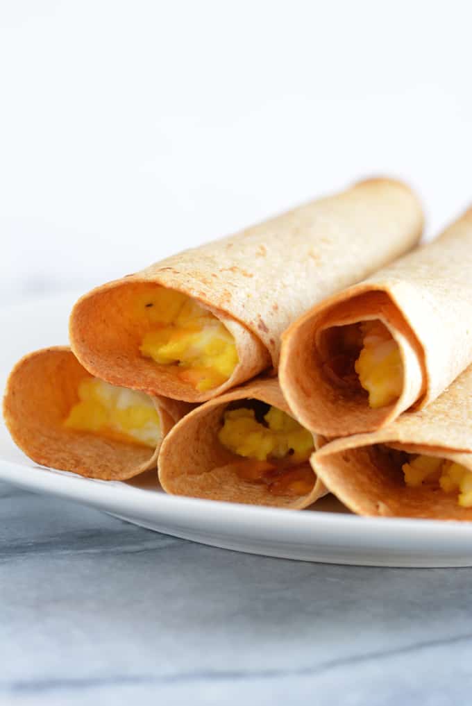 Breakfast taquitos filled with eggs, ham and cheese stacked on a white plate.