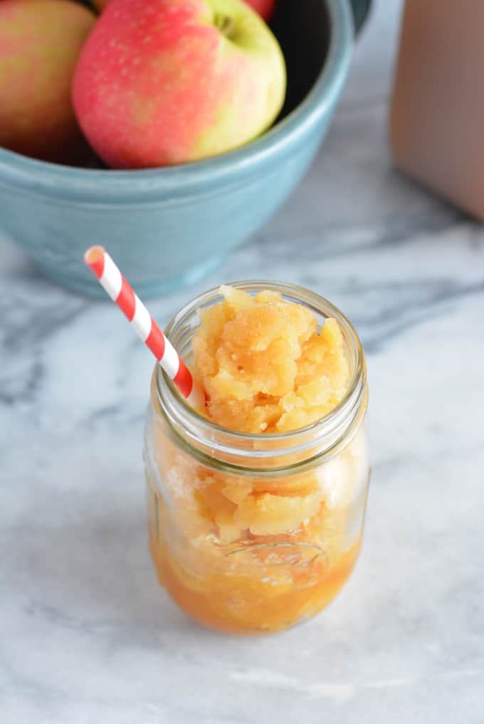 Frozen Apple Cider Slushy in a mason jar with a white and red striped straw next to a bowl of apples.