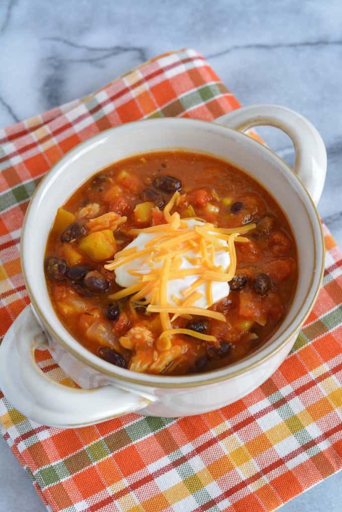 A bowl of turkey chili with cheddar cheese and sour cream on top place on a orange plaid napkin.