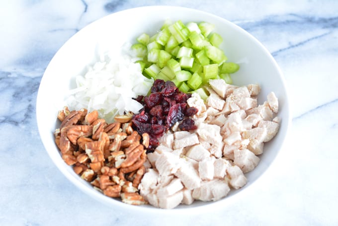 Diced chicken, chopped celery, chopped onion, pecans, and dried cranberries in a white bowl.