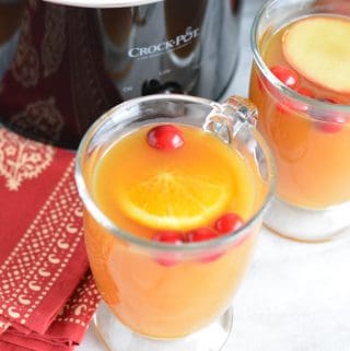 Apple cider in a glass mug in front of a black slow cooker.