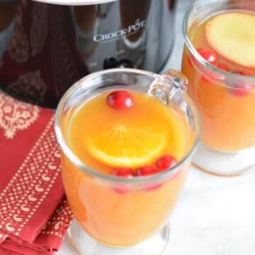 Apple cider in a glass mug in front of a black slow cooker.