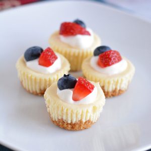 Mini cheesecakes with whipped cream, strawberries, and blueberries on a white plate.